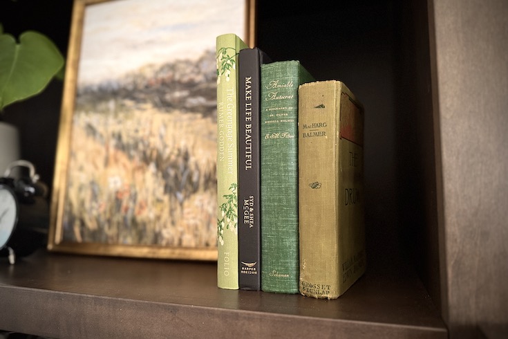 Four books all in different shades of green placed on shelf next to an artwork.