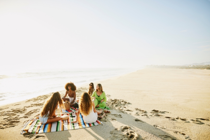 Wide shot of smiling female friends relaxing on blanket on tropical beach at sunset while on vacation.