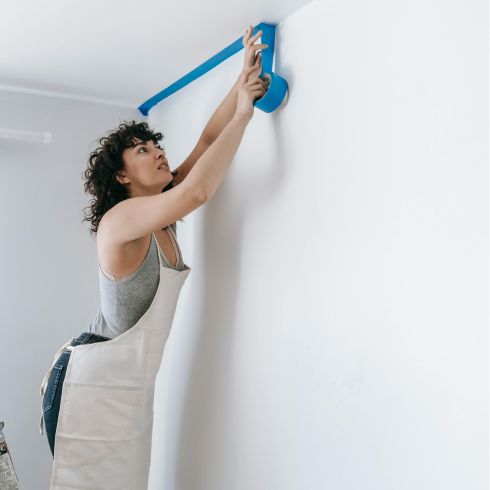 A person standing on a ladder with an apron on as they add painter's tape to a wall.