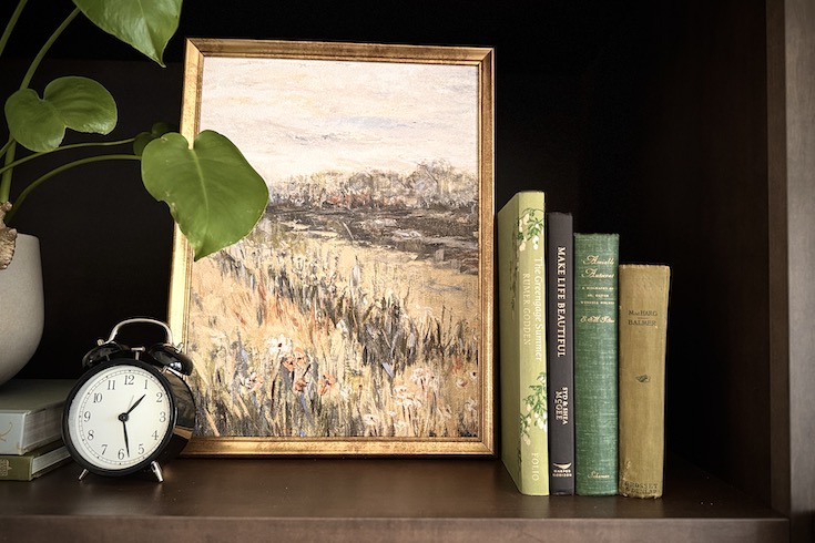 Artwork of landscape in between a plant and books on shelf.