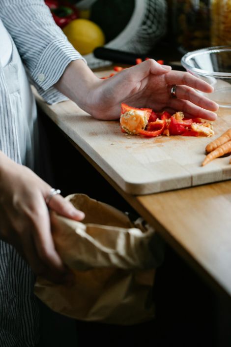 A persons hand drags red pepper peels over a cutting board and into a paper bag. 