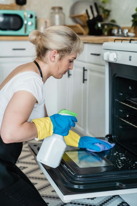A woman in a white t-shirt holding a white and green plastic spray bottle cleaning the oven.