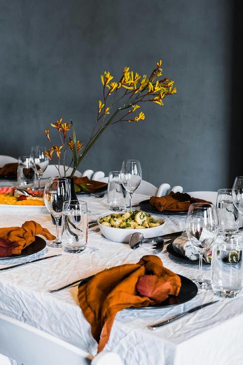 Elegant Table Decoration with yellow flowers in a vase, orange napkins, dark dishes, white table cloth and a grey wall