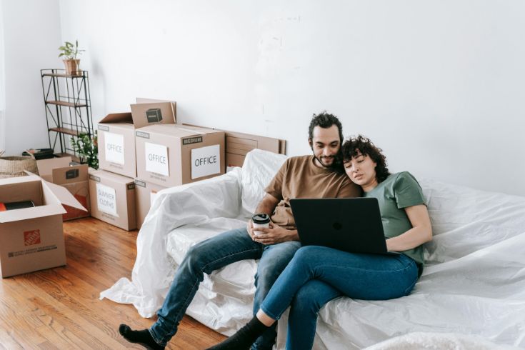 A couple is sitting on their couch in an apartment with boxes around them. 