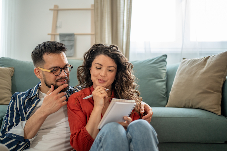 Young couple sitting comfortably on the floor beside the sofa, happily taking notes and planning their home finances together, discussing budgets, savings, and future investments