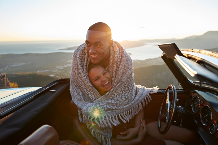 Happy man embracing girlfriend from behind while wrapped in blanket on road trip.