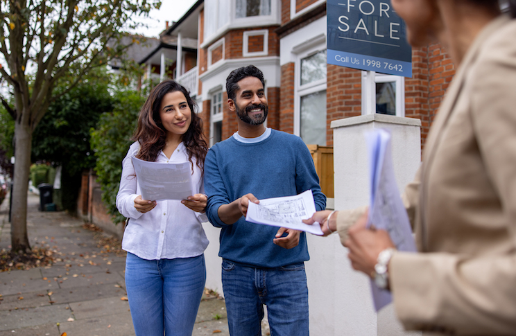 Real estate agent handling the blueprints of a house for sale to an Indian couple on the street and smiling
