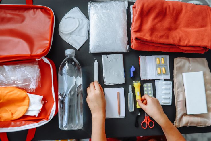 Close-up of women's hands packing a flashlight in doomsday prepping on the table at home. Includes warm clothing, flashlight, power bank, thermal blanket, medicines, water, and other emergency supplies.