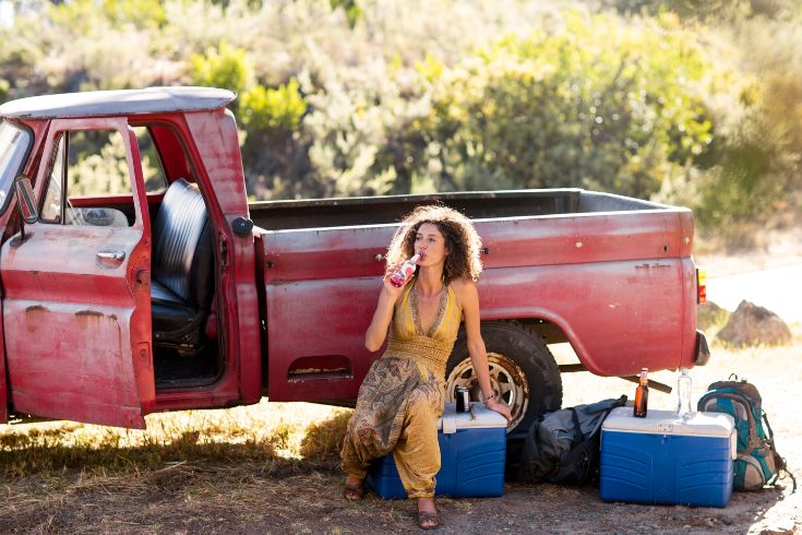 Young woman sitting next to old rusty pick up truck drinking from bottle.