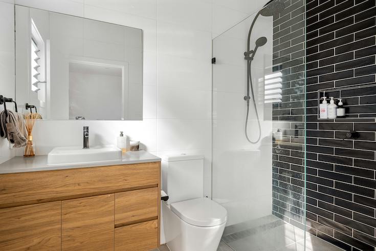 Interior of a Modern, Minimalist Bathroom with a white toilet, walk-in shower with black tiles, wooden vanity and white sink