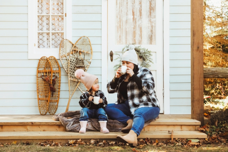 A beautiful little girl, three years old, smiles as she sits on a charming, rustic porch, with her mom. They are enjoying mugs of hot chocolate.