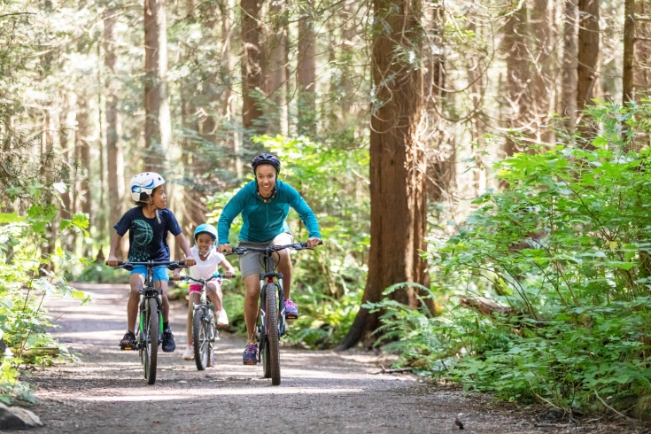 A woman and her two kids ride bikes in the forest in the sunshine.