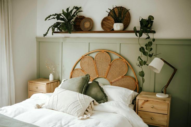 Bedroom with green wainscotting, a rattan headboard, two wooden bedside tables, and wall shelf with plants and woven plates