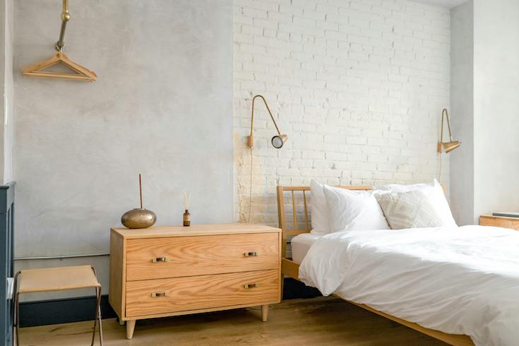 Bedroom with simple golden wall lamps on a textured grey wall with a wooden dresser and white bed linens