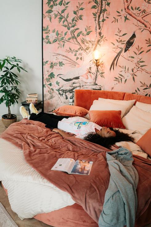 Woman Lying on Her Bed with large framed fabric artwork above the bed covered in red and orange linens and cushions