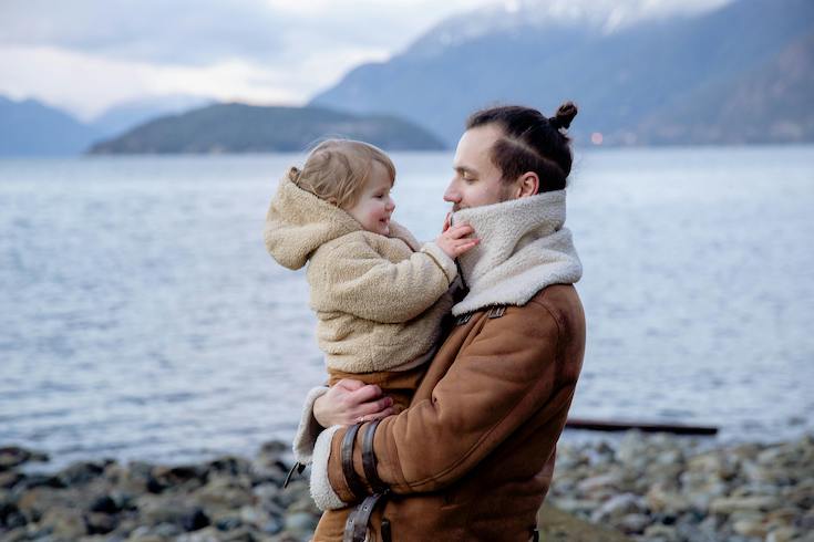 Loving father holding little kid on hands while standing near water on stony coast near Victoria, BC